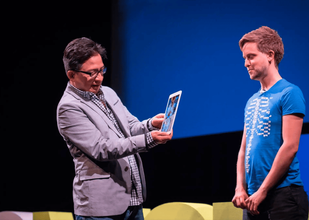 An image from a conference presentation featuring two individuals on stage. The person on the left, dressed in a grey blazer and glasses, is animatedly holding a tablet and looking at its screen. The individual on the right, wearing a blue T-shirt with a digital design, is standing attentively with his hands together, facing the first speaker but looking slightly downwards. They are both standing in front of a simple set with a plain blue backdrop, indicating a focus on the interaction and the digital device.