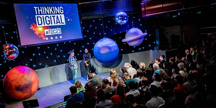 An image from the Thinking Digital Conference, displaying a vibrant stage setting with a space theme. Two speakers stand on the stage, with one appearing to address the audience. The backdrop features a large screen with the conference branding, "#TDC23", signifying the event's year. Above the crowd, there are large 3D models of planets suspended from the ceiling, adding to the cosmic ambiance. The audience is seated in rows, attentively watching the presentation. The venue has a mix of warm and cool lighting, giving it a dynamic and engaging atmosphere.