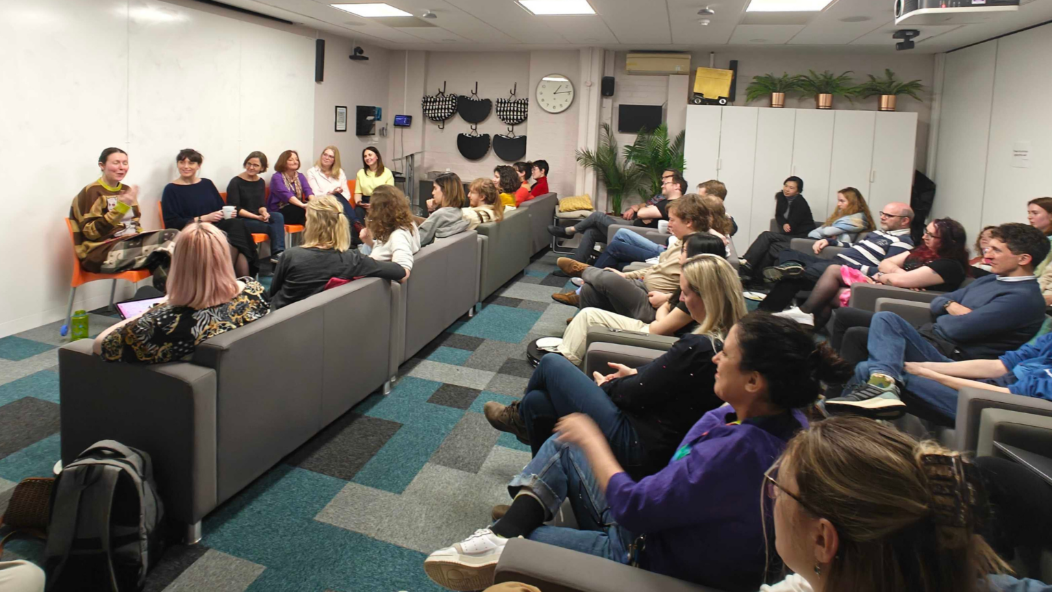 A panel discussion taking place in a modern, casual office environment. A diverse group of approximately 30 people sits in rows of soft, modular seating. They are attentively facing two women who are seated at the front of the room. One woman, speaking, holds papers and a mug, while the other listens. The room has a relaxed vibe, with a mix of standing lamps and plants, a clock on the wall reading ten past ten, and coats hung neatly on a rack in the background.