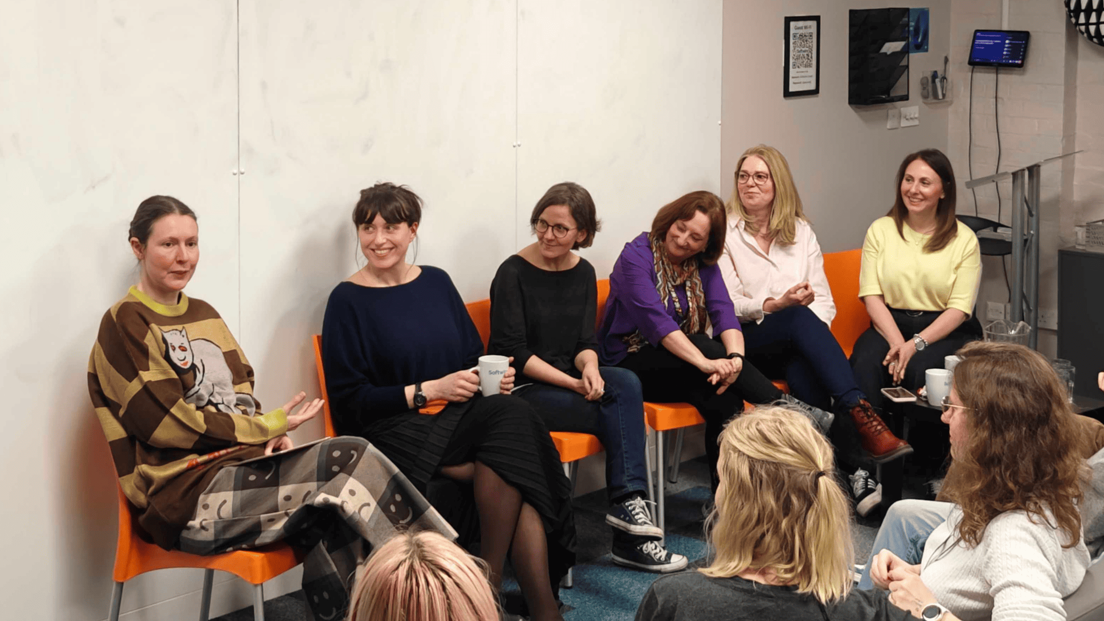A panel discussion taking place in a modern, casual office environment. A diverse group of approximately 30 people sits in rows of soft, modular seating. They are attentively facing two women who are seated at the front of the room. One woman, speaking, holds papers and a mug, while the other listens. The room has a relaxed vibe, with a mix of standing lamps and plants, a clock on the wall reading ten past ten, and coats hung neatly on a rack in the background.