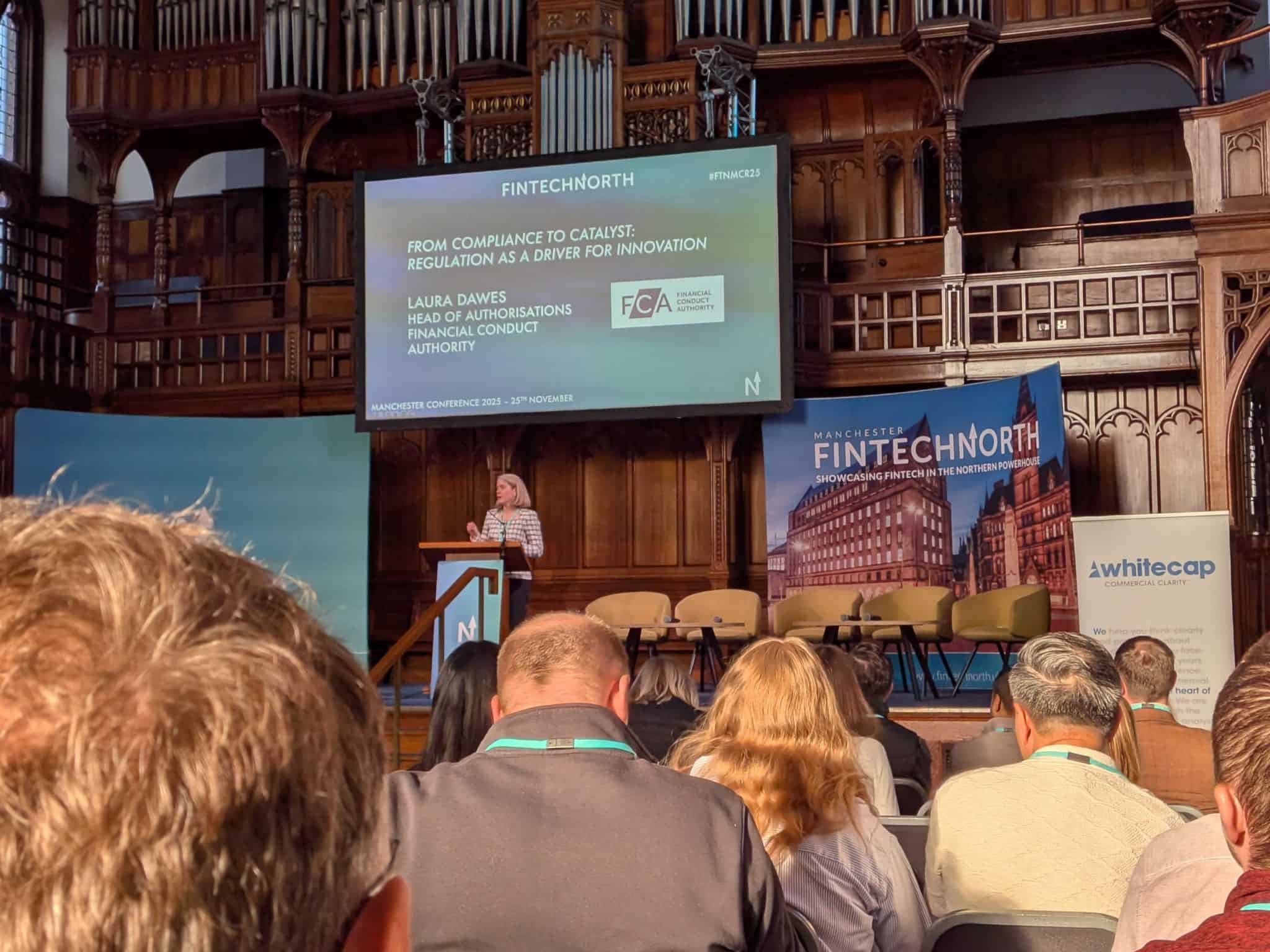 A speaker stands at a lectern delivering a talk at the Manchester FinTech North Conference. The presentation slide behind her reads “From Compliance to Catalyst: Regulation as a Driver for Innovation” by Laura Dawes from the Financial Conduct Authority. The venue features ornate wooden architecture and an organ. The audience sits facing the stage, with FinTech North and Whitecap banners visible.