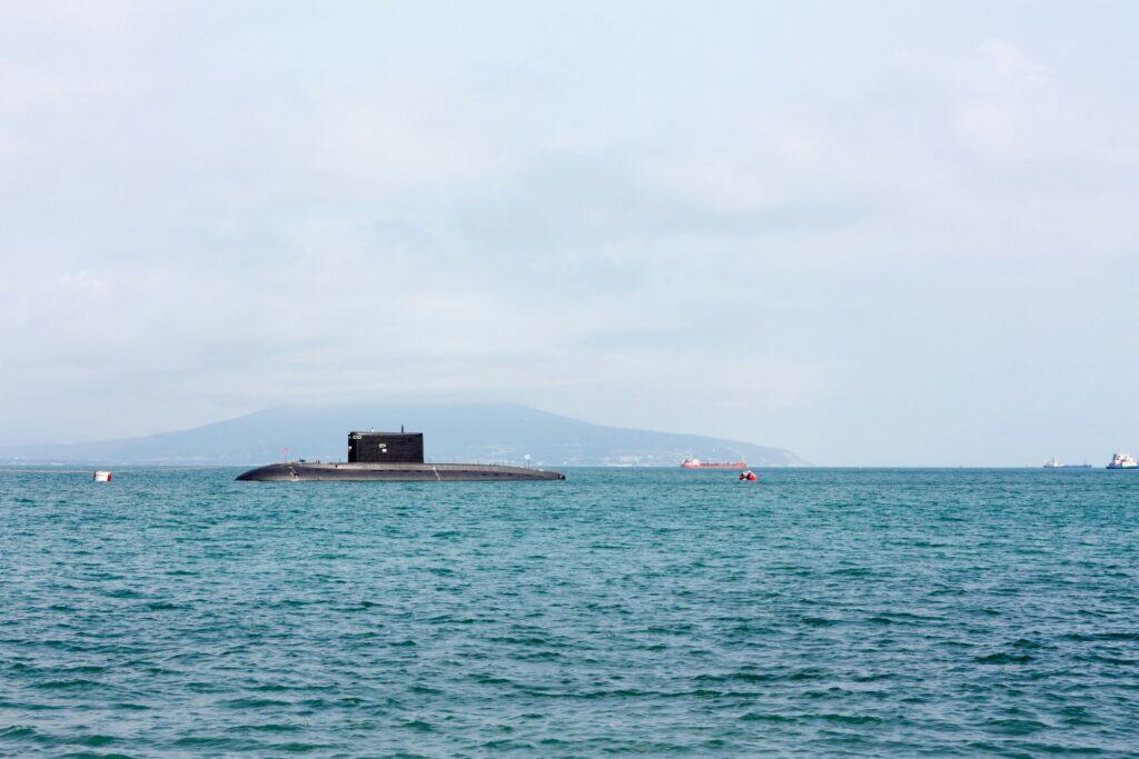 A submarine surfaced on calm open water, with distant ships and a hazy coastline visible on the horizon under a pale sky.