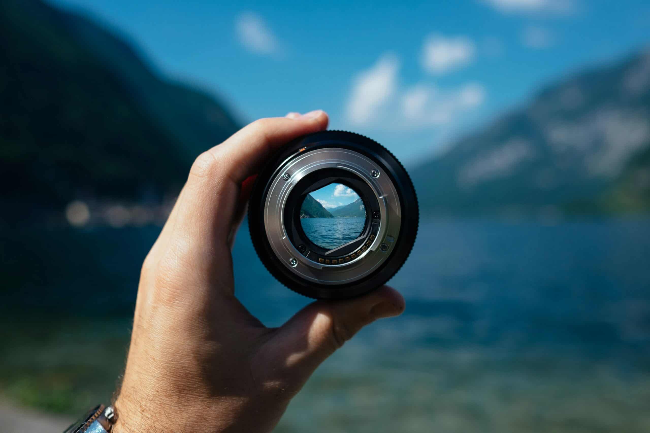 Hand holding a camera lens up to a mountain lake, with the landscape sharply framed through the lens while the background remains softly blurred.