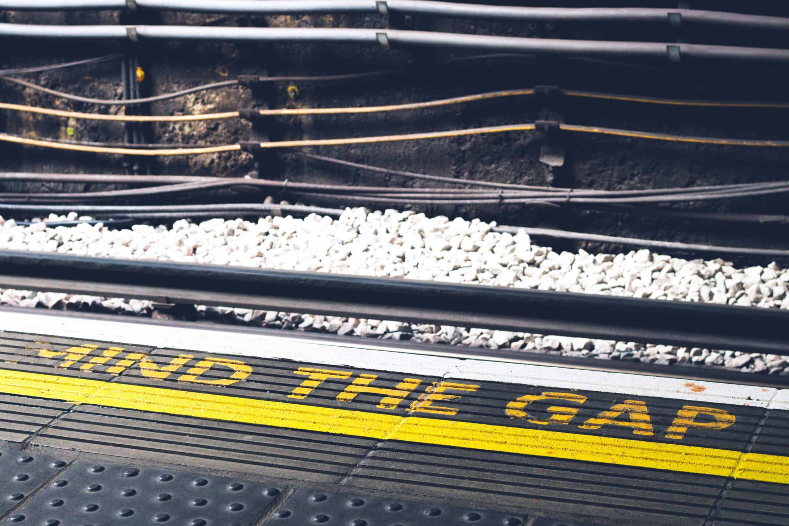 A railway platform edge with a yellow safety line and the words “Mind the gap” painted on the ground, with train tracks, gravel, and cables visible beyond the platform.