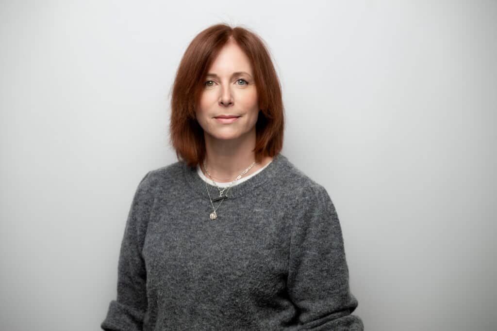 Portrait of a woman with shoulder-length red hair, wearing a gray sweater, looking at the camera against a light gray background.